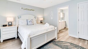 A bedroom with a white bed, two bedside tables with lamps, a ceiling fan, and a door leading to a bathroom.