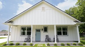 A white single-story building with a gabled roof, a front porch with two chairs, and a blue door. - Modern Nest - Brand New House w/ modern amenities (Bryan)