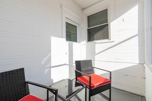 En balkon med to stole og et glasbord, hvide vægge og to vinduer med skodder.