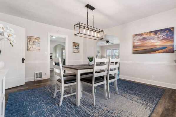 A dining area with a rectangular table, white chairs, a blue rug, and a ceiling fan.