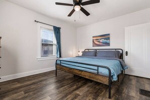 A bedroom with a bed, a ceiling fan, a window with curtains, a door, and a painting on the wall. - Larger 2 Bedroom Apt in the Heart of Canton, Minutes From HOF and Both Hospitals (Canton)