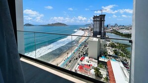 Vista da un balcone su una spiaggia, con cielo sereno, un'isola in lontananza e uno skyline con edifici e una gru edile.
