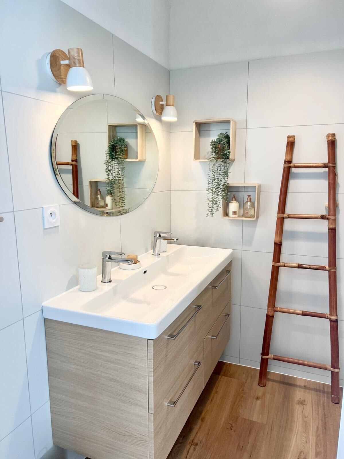 A bathroom with a large mirror, a sink, and two wall-mounted lights. There are wooden shelves with plants and toiletries. A wooden ladder is leaning against the wall.