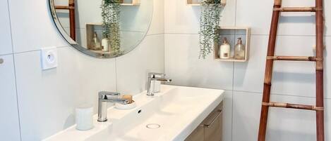 A bathroom with a large mirror, a sink, and two wall-mounted lights. There are wooden shelves with plants and toiletries. A wooden ladder is leaning against the wall.