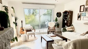 Una sala de estar acogedora con un sofá blanco, una mesita de centro, una guitarra y una silla. Hay un ventanal grande con vistas a la vegetación.