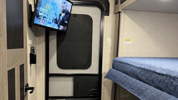 Un intérieur de camping-car compact, doté d’un téléviseur fixé au mur, de lits superposés et d’une porte avec un panneau indiquant la sortie.