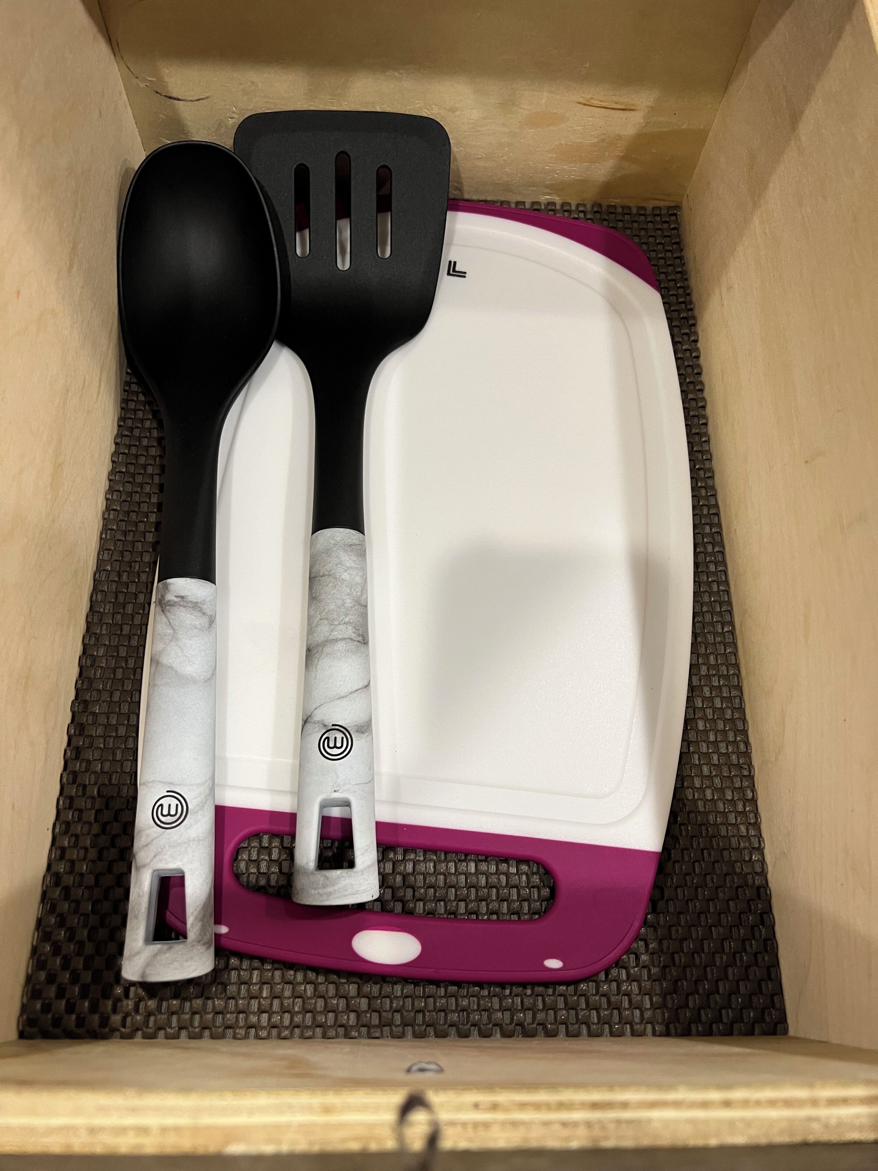 A drawer containing a white and purple cutting board, a black and white spatula, and a black ladle.