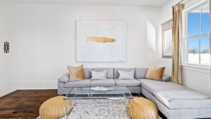 A modern living room with a grey sectional sofa, a glass coffee table, and a large abstract painting on the wall. - Pristine 4BD w Pool | Driveway Parking | Balcony (New Orleans)