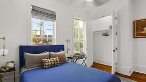 A bedroom with a large bed, two windows, a door leading to a closet, and a small table with books. - Pristine 4BD w Pool | Driveway Parking | Balcony (New Orleans)