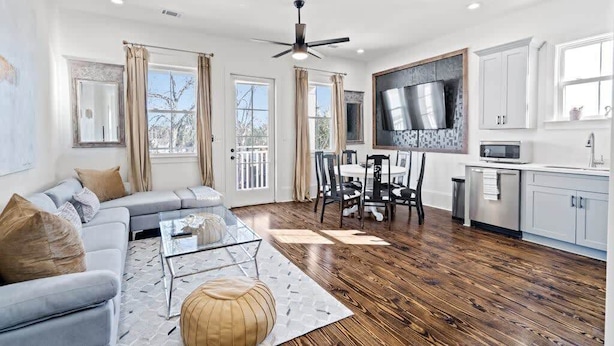 A modern living room with a sofa, coffee table, and dining area. There is a kitchen with white cabinets and a large TV. - Pristine 4BD w Pool | Driveway Parking | Balcony (New Orleans)