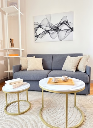 Un salon moderne avec un canapé gris, deux tables rondes, une table basse en marbre, une bibliothèque et une œuvre d’art murale.