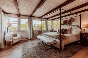 A four-poster bed with a bench, a sofa, and a chair in a room with large windows and a view of trees.