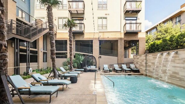 A pool area with lounge chairs, a palm tree, and a building with balconies. - Charming 1,100 sq. ft. apt (Plano)