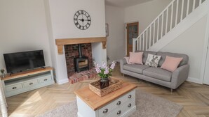 A living room with a TV, sofa, fireplace, staircase, and a clock on the wall. - The Miners Cottage (Berwick-upon-Tweed)