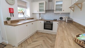 A modern kitchen with white cabinets, a built-in oven, a microwave, and a toaster. There is a clock on the wall and a fire blanket below it. - The Miners Cottage (Berwick-upon-Tweed)