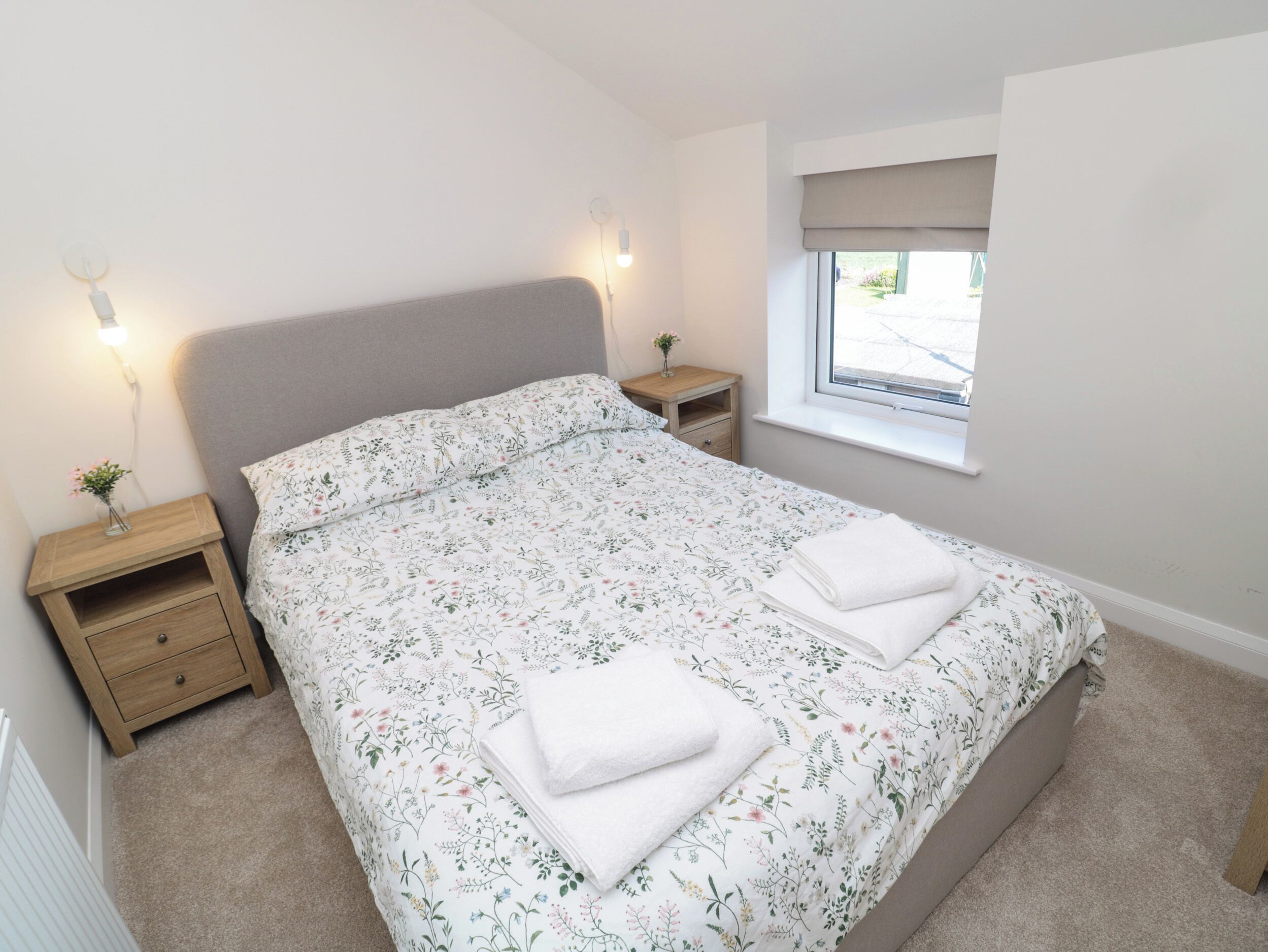 A bedroom with a large bed, two bedside tables, a window with a view, and a nightstand with a vase.