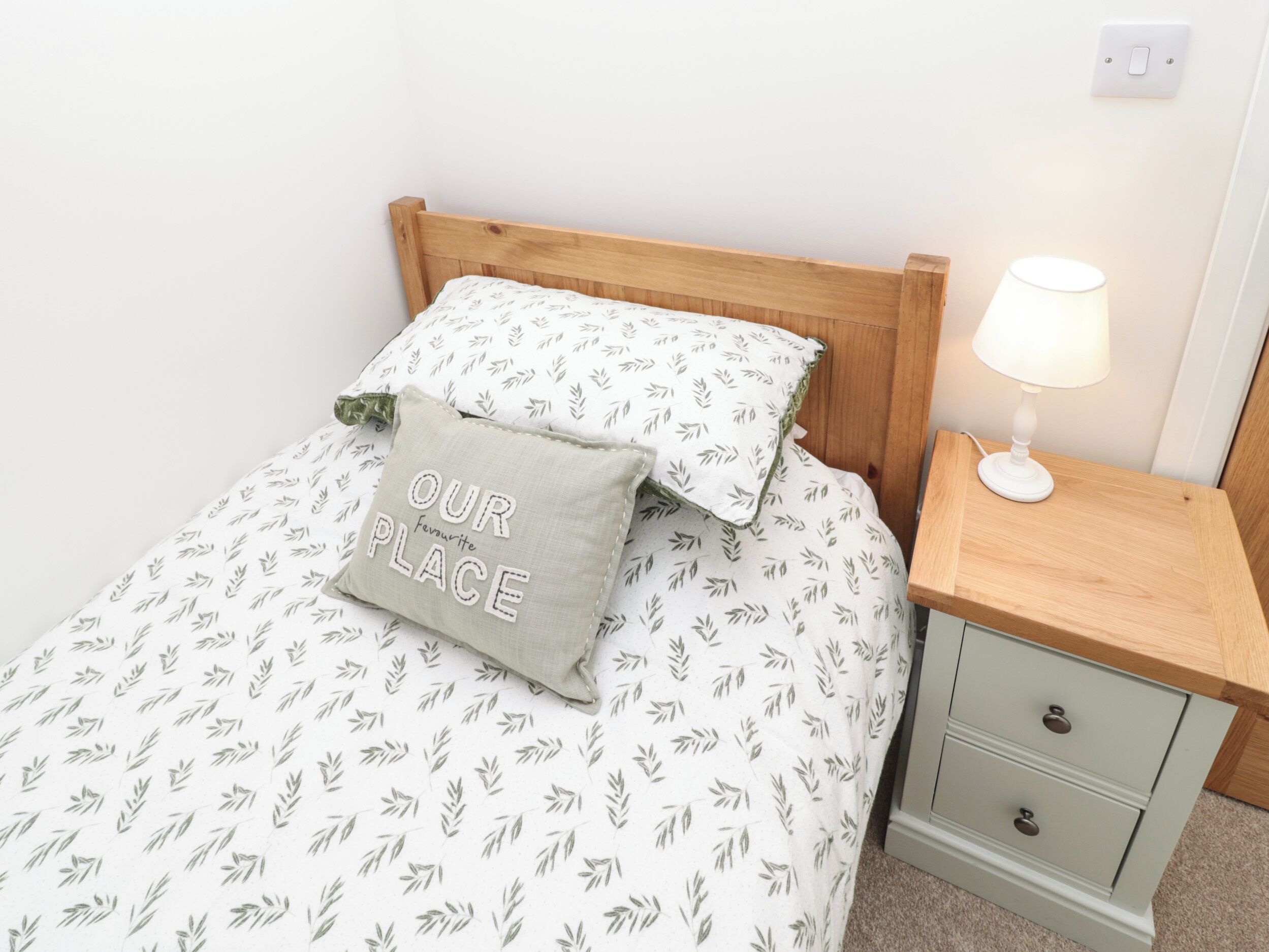 A neatly made bed with a leaf-patterned bedspread and pillows, including one with "OUR PLACE" text. There's a bedside table with a lamp and a drawer.