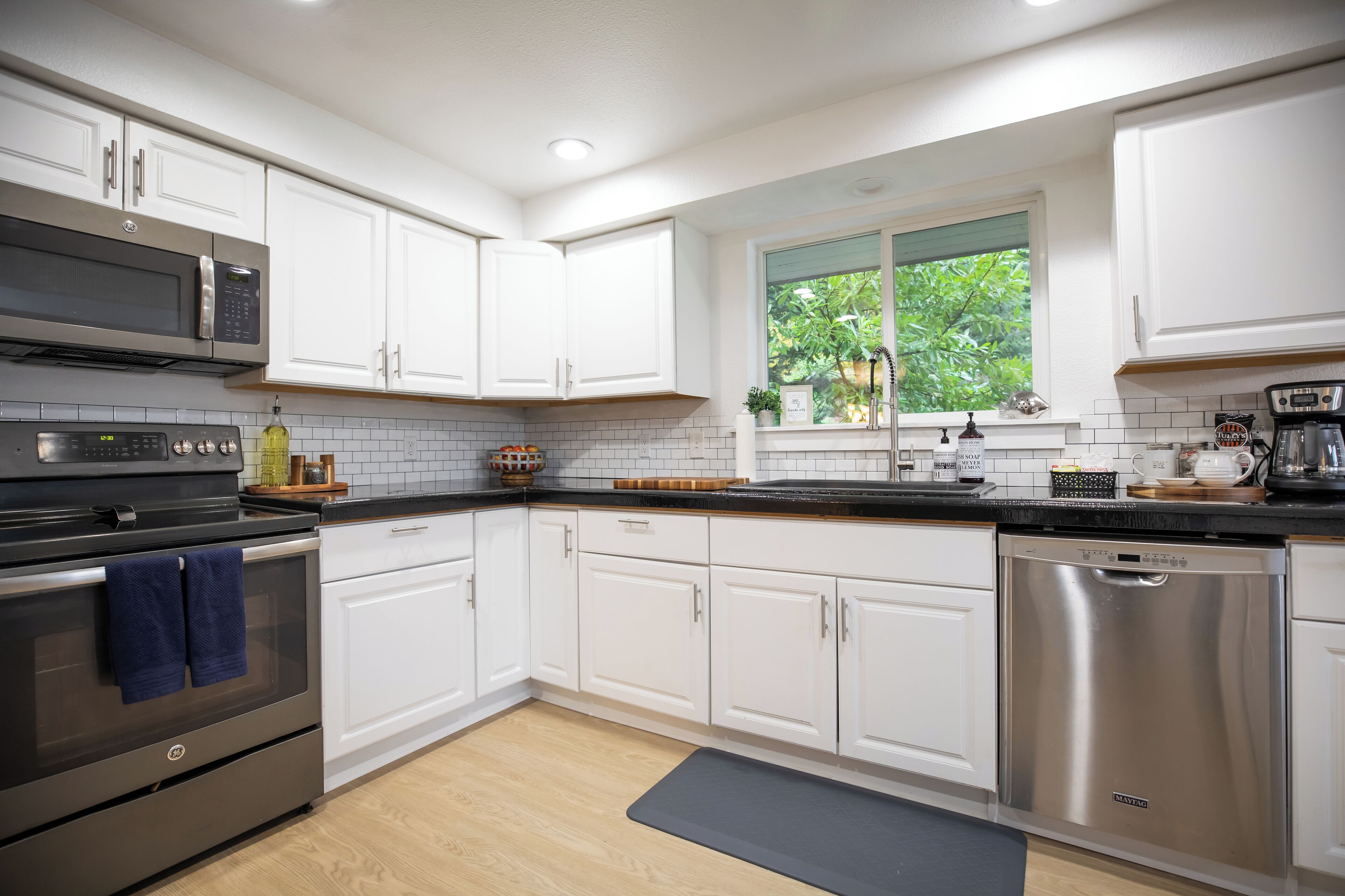 A modern kitchen with white cabinets, stainless steel appliances, and a window with a view of greenery.