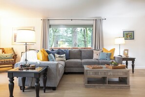 A living room with a grey sectional sofa, two side tables, a wooden coffee table, and a large window with curtains. - Lakeside home on Horseshoe Lake: kayaks, paddle boards, fire pit, games, fishing (Port Orchard)