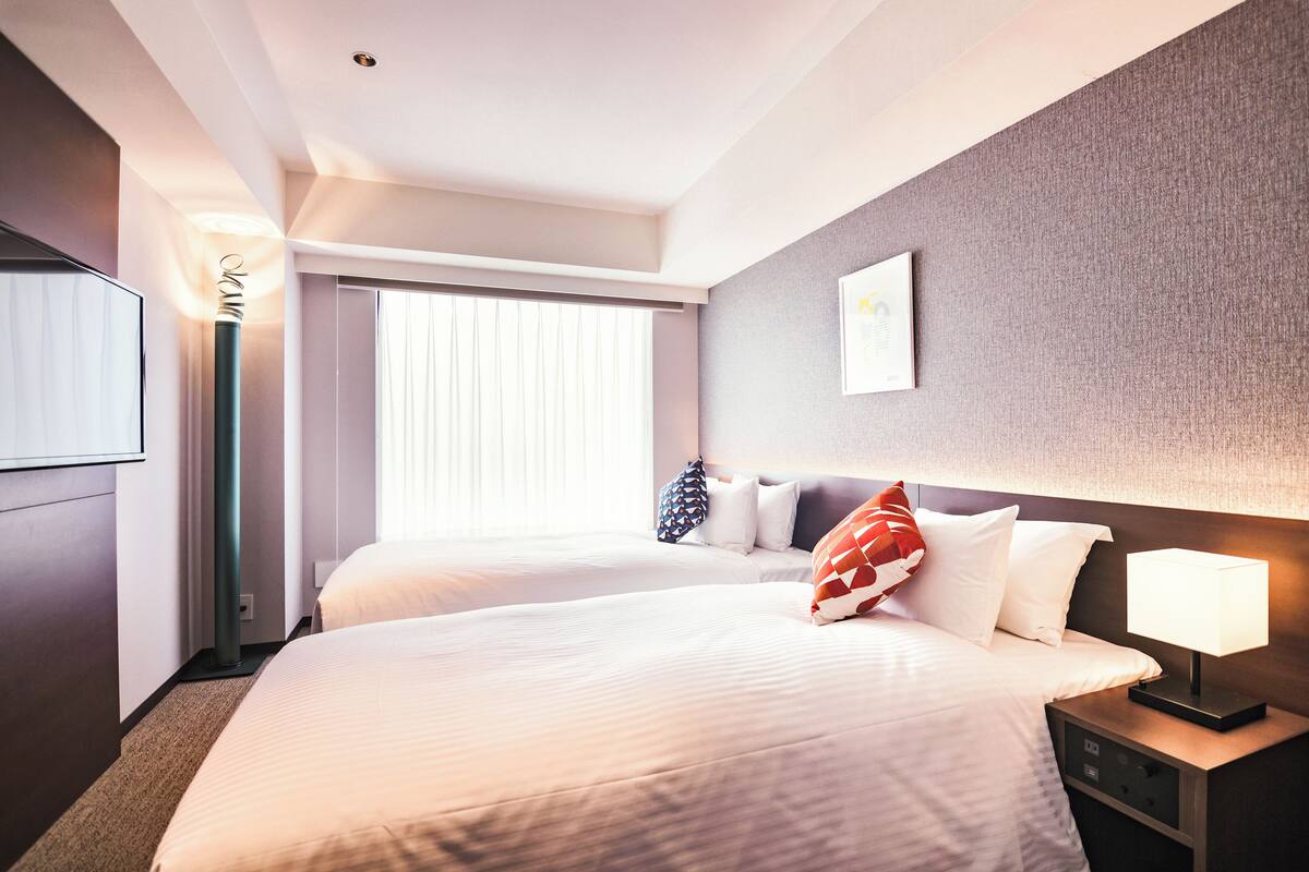 A hotel room with two beds, a bedside table with a lamp, a wall-mounted TV, and a window with blinds.