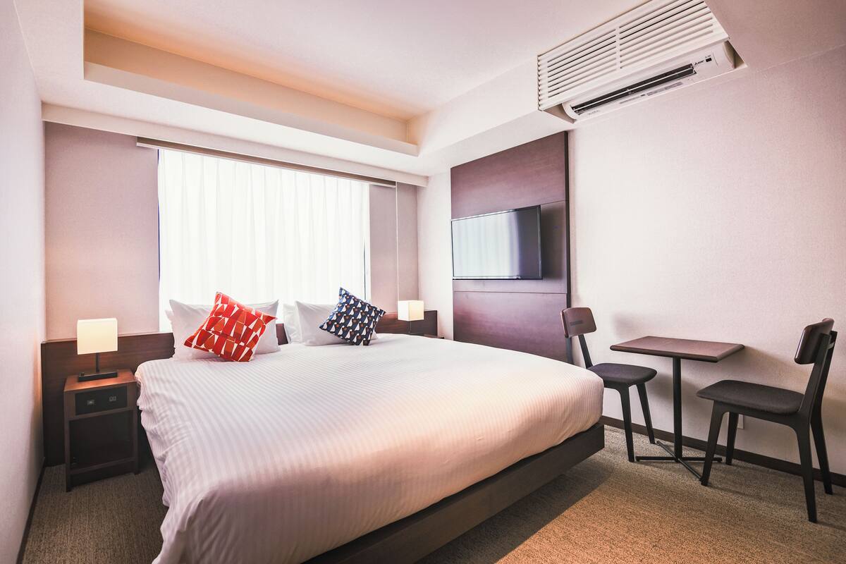 A hotel room with a large bed, a TV mounted on the wall, a desk with two chairs, and an air conditioning unit.