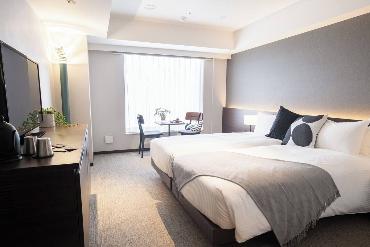 A modern hotel room with a large bed, a desk with a coffee maker, a chair, and a window with blinds.