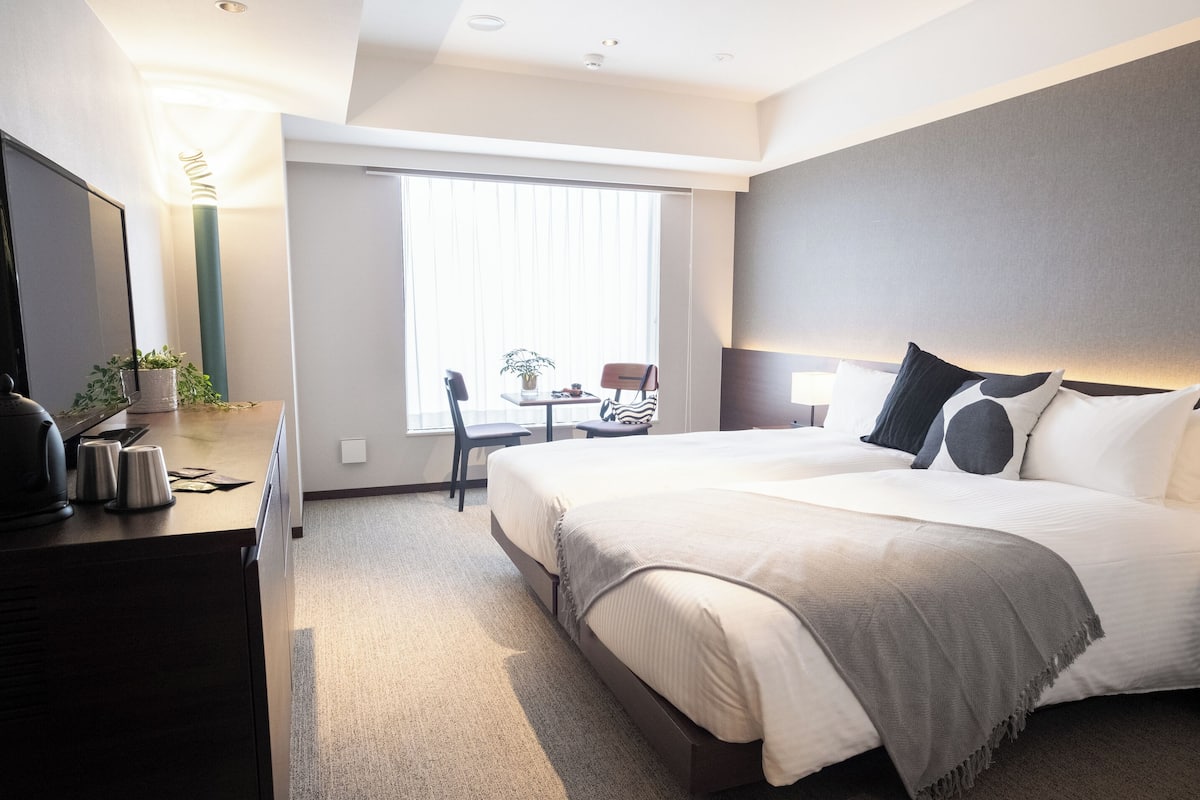 A modern hotel room with a large bed, a desk with a coffee maker, a chair, and a window with blinds.