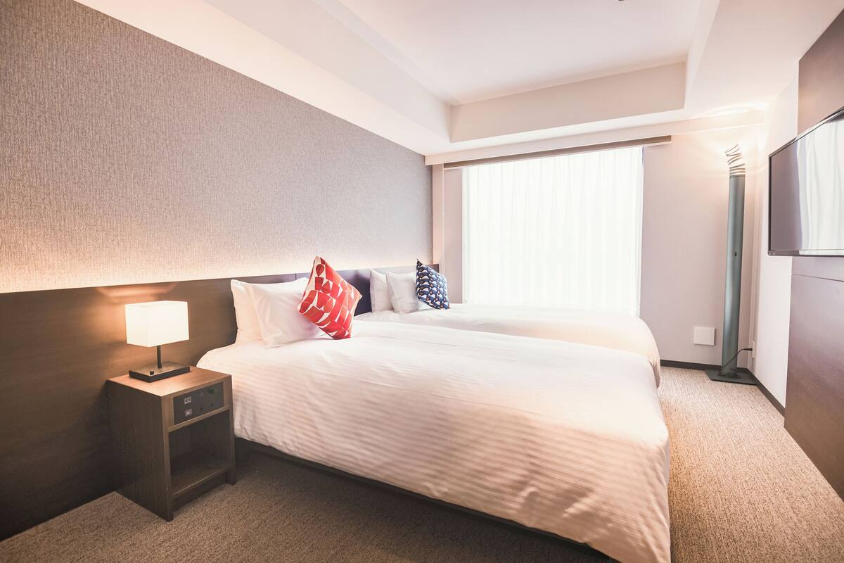 A hotel room with a large bed, a nightstand with a lamp, and a large window with blinds.