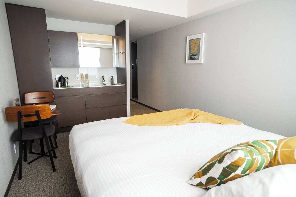 A hotel room with a bed, a desk with a chair, a mirror, a sink, and a framed picture on the wall.