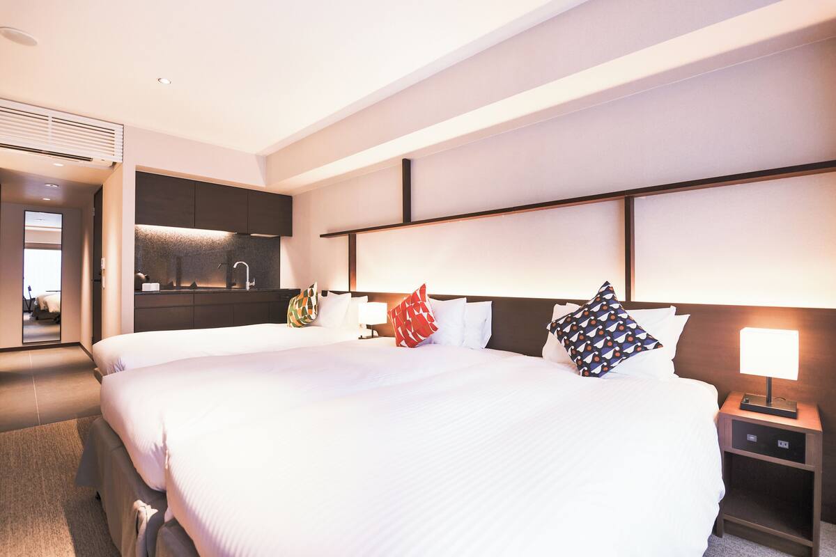A hotel room with two beds, a small desk, a nightstand, a mirror, and a kitchenette.