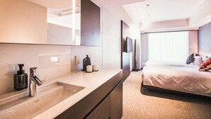 Una habitación de hotel moderna con un lavamanos grande en el baño, un espejo, una cama con televisor y una ventana con persianas.