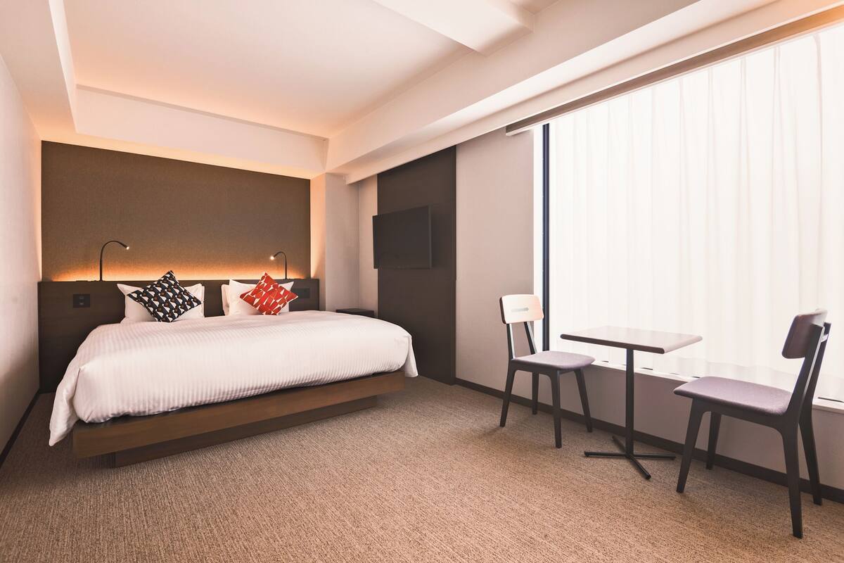 A modern hotel room with a bed, a desk, two chairs, a TV, and a large window with blinds.
