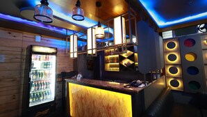 A bar area with a neon-lit bar counter, a refrigerated beverage dispenser, and a seating area with a wooden wall and blue ceiling accents. - HOTEL JAI HARSH BY DIVYANSHI HOSPITALITY (Jaipur)