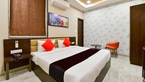 A hotel room with a large bed, a bedside table with a phone, an air conditioning unit, a colorful painting on the wall, and a small table with a chair. - HOTEL JAI HARSH BY DIVYANSHI HOSPITALITY (Jaipur)