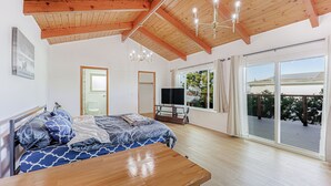 Un dormitorio con techo de madera, cama con ropa de cama estampada, mesita de noche, televisor y puerta corrediza de vidrio a una terraza exterior.