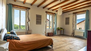 A bedroom with a wooden floor, a bed with a brown bedspread, and large windows with curtains. - Maison Entière Haut Valromey (HAUT-VALROMEY)