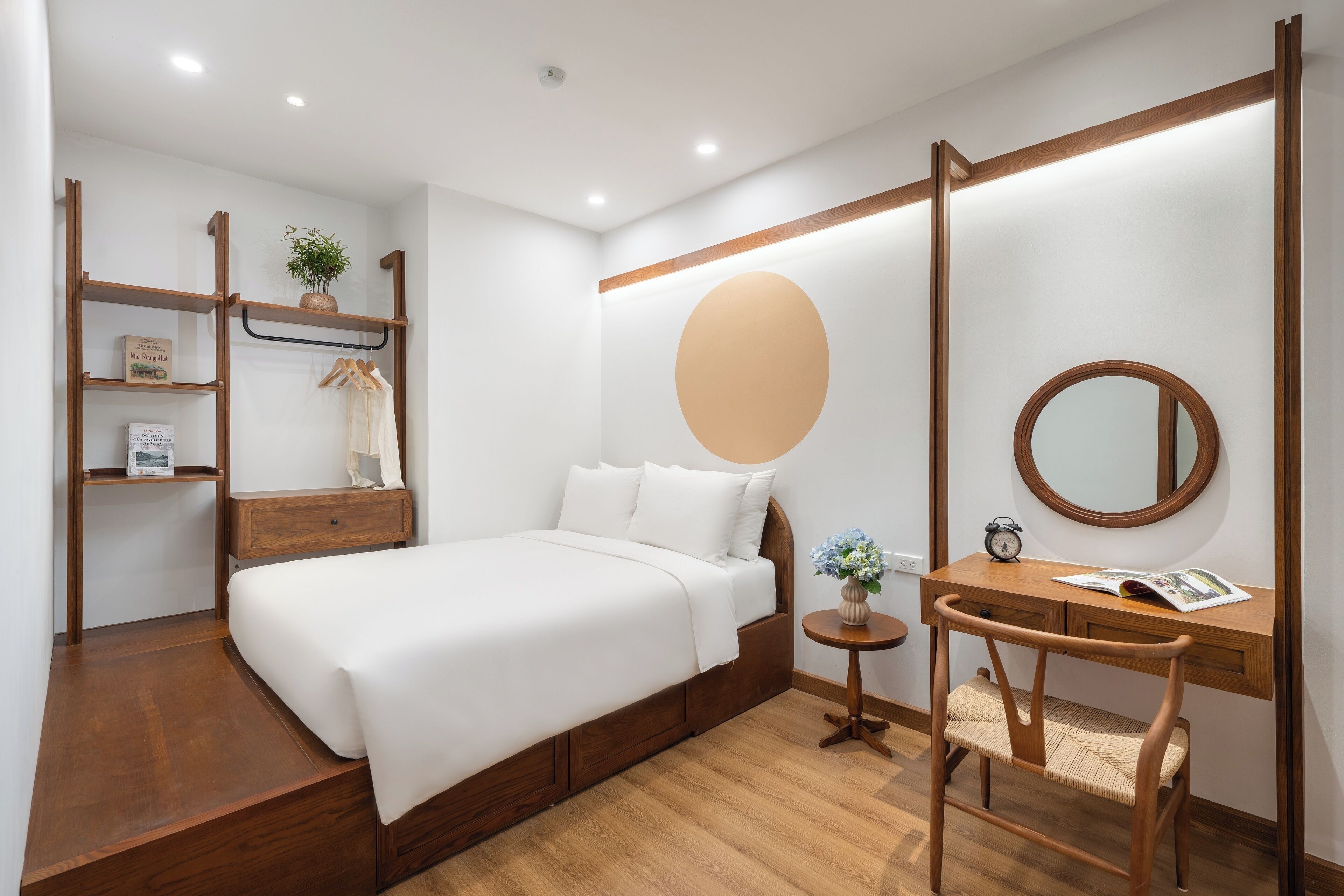 A hotel room with a wooden bed, a desk with a chair, a round mirror, and a shelf with a plant.