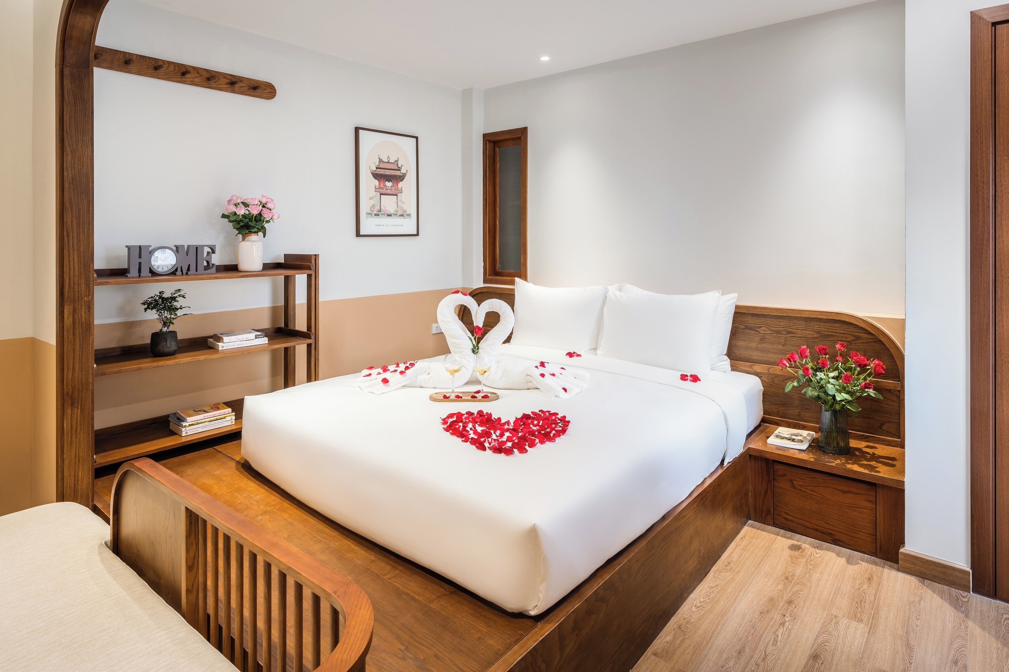 Ein Hotelzimmer mit einem ordentlich bezogenen Bett, einem hölzernen Kopfteil, einem Nachttisch mit Blumenvase sowie einem Regal mit Büchern und einer Uhr.