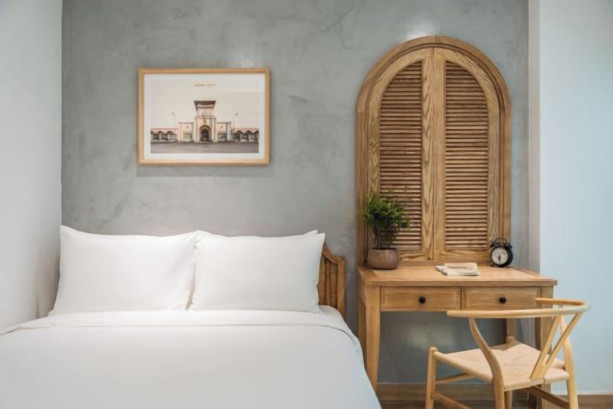 A bedroom with a bed, a wooden desk with a chair, a framed picture on the wall, and a wooden arched window.
