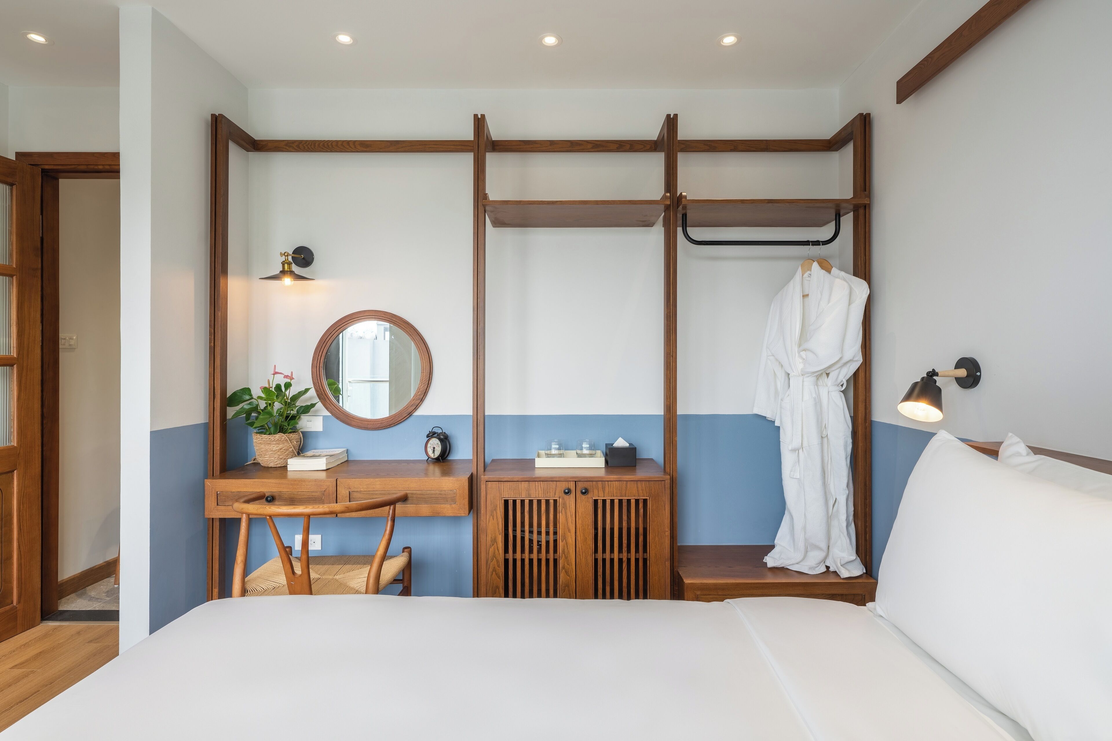 A bedroom with a bed, a wooden desk, a mirror, a hanging robe, and a bedside lamp.