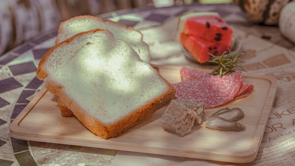 Slices of white bread on a wooden board with a piece of salami and a slice of watermelon on a table with a newspaper underneath. - Le Parfum Apartment And Hotel (Hanoi)