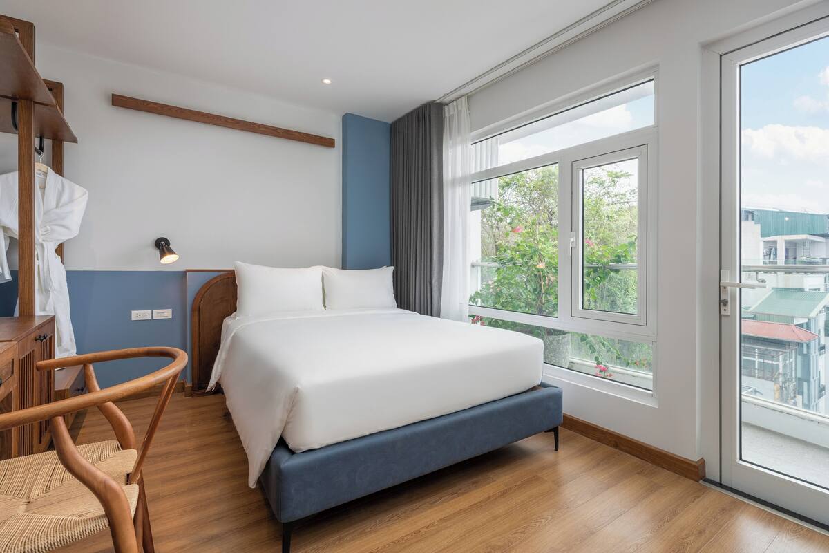 Une chambre d’hôtel moderne avec un grand lit, une chaise en bois, un bureau avec une lampe et une vue sur la verdure par la fenêtre.