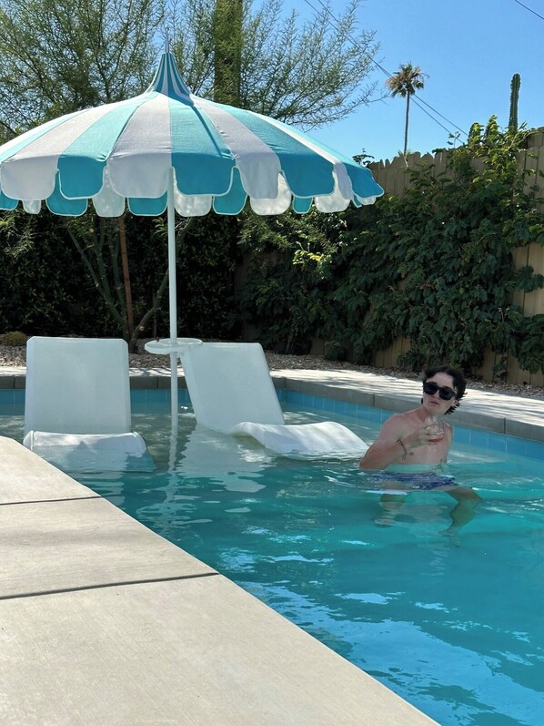 Una piscina con un paraguas azul y blanco a rayas, dos sillas reclinables blancas y una persona en el agua.