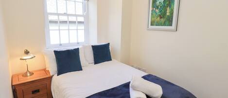 Una cama bien hecha con edredón blanco y azul marino, una mesita de noche de madera con una lámpara, una ventana con un visillo floral y un cuadro en la pared.