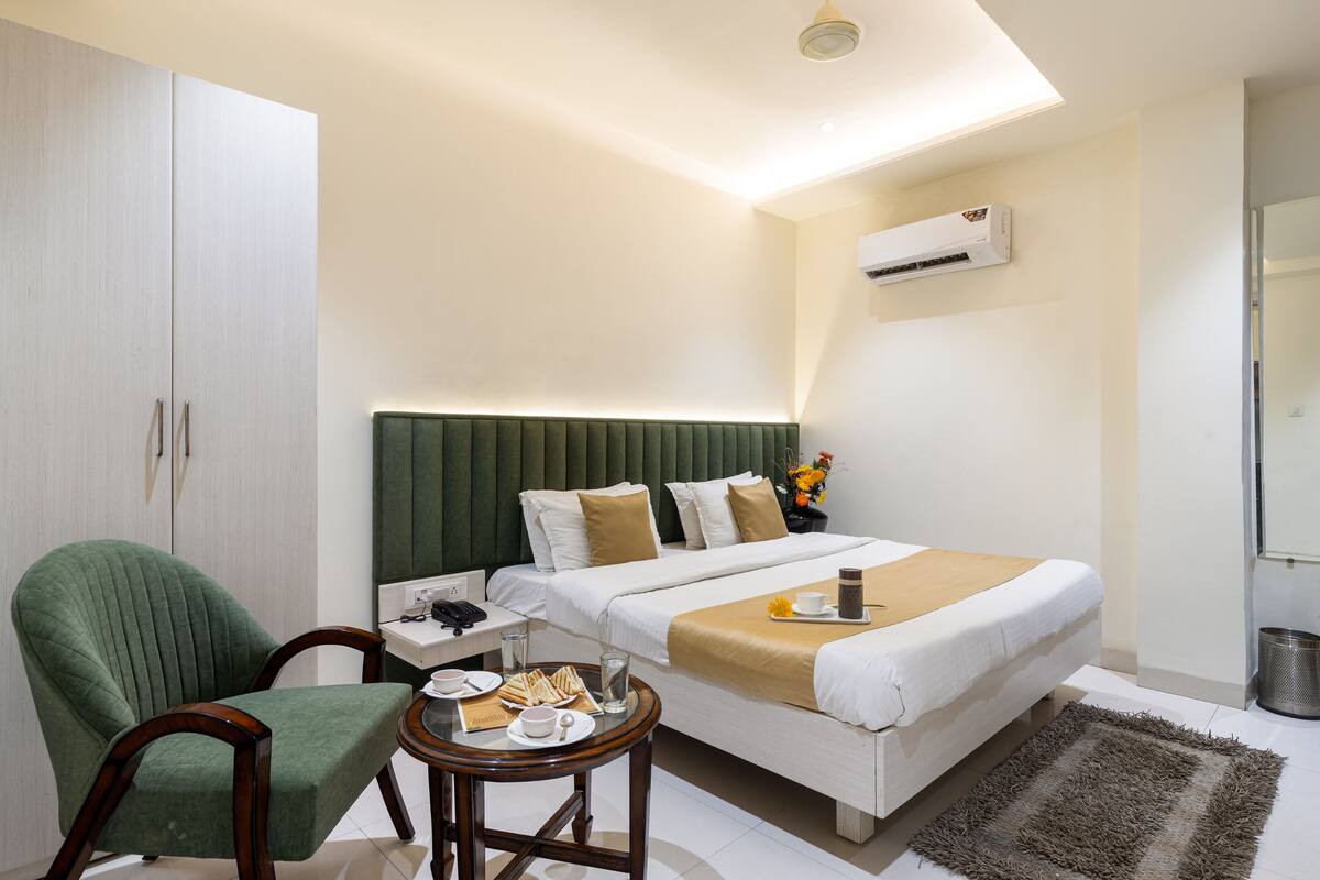 Una habitación de hotel moderna con una cama, un sillón verde, una mesita redonda con cosas para el desayuno y una mesita de noche con una lámpara.