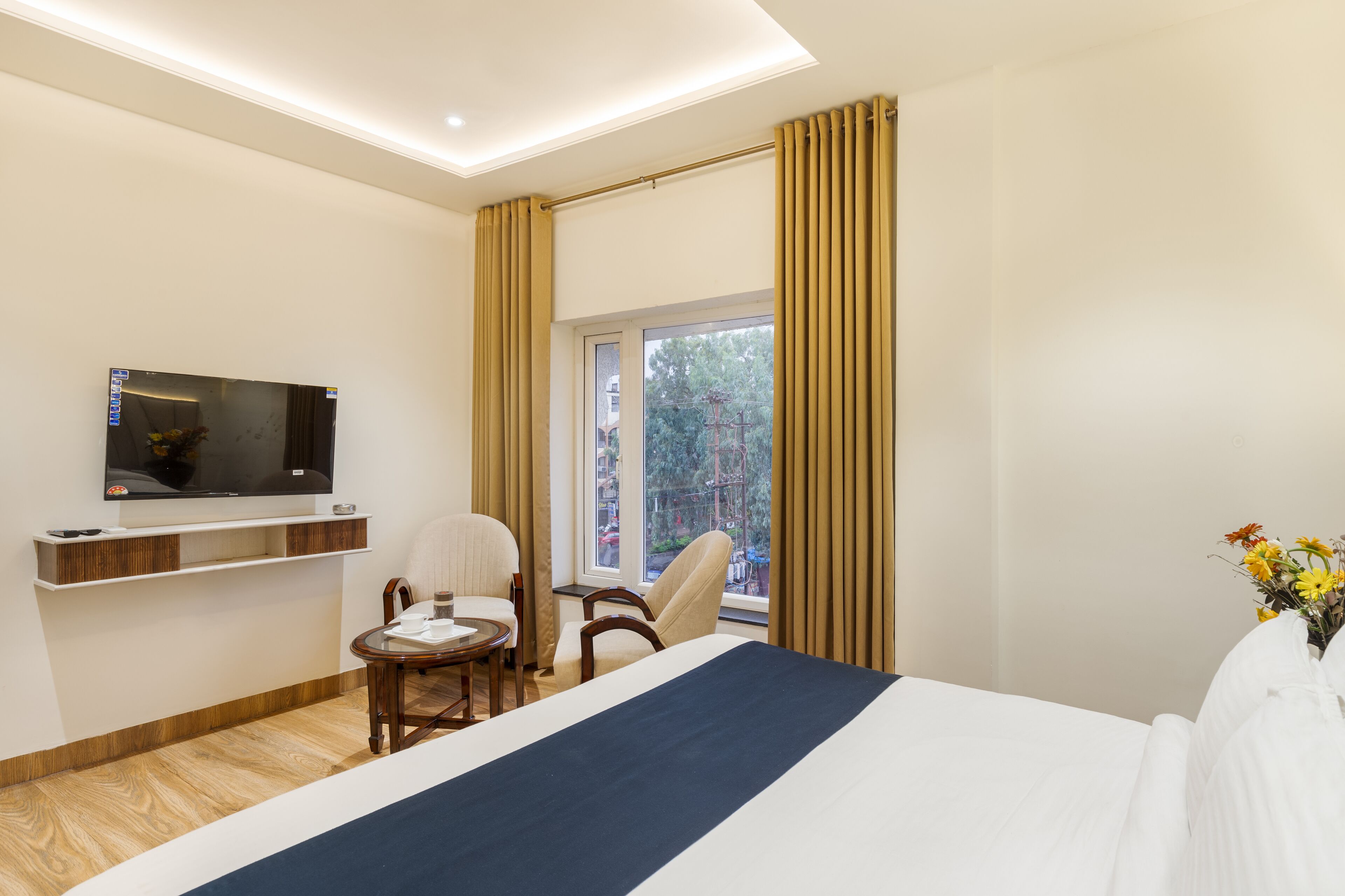 A hotel room with a bed, a TV mounted on the wall, a small table with a chair, and a window with curtains.