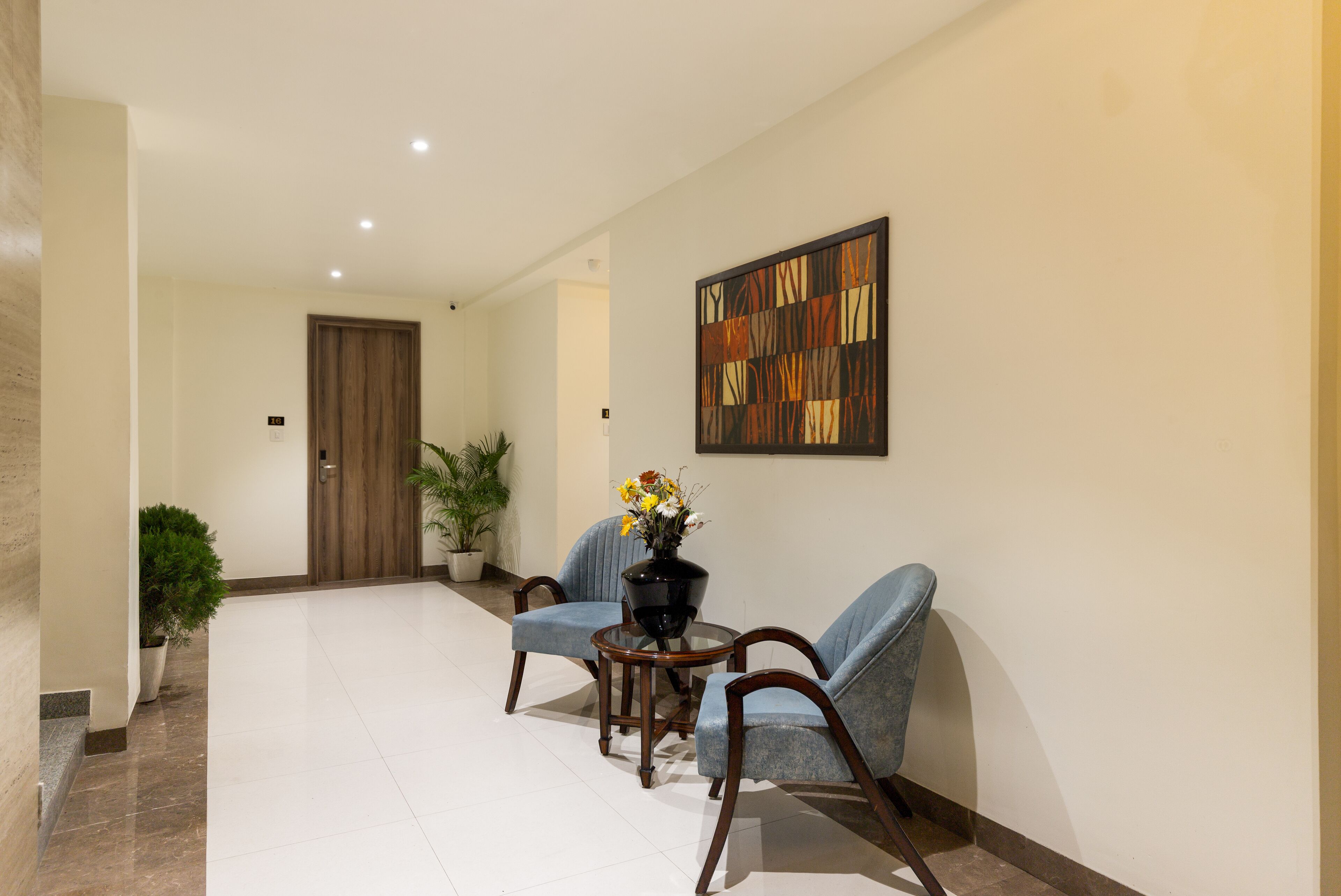 A hallway with two blue armchairs, a small table with a vase of flowers, and a large abstract painting on the wall.