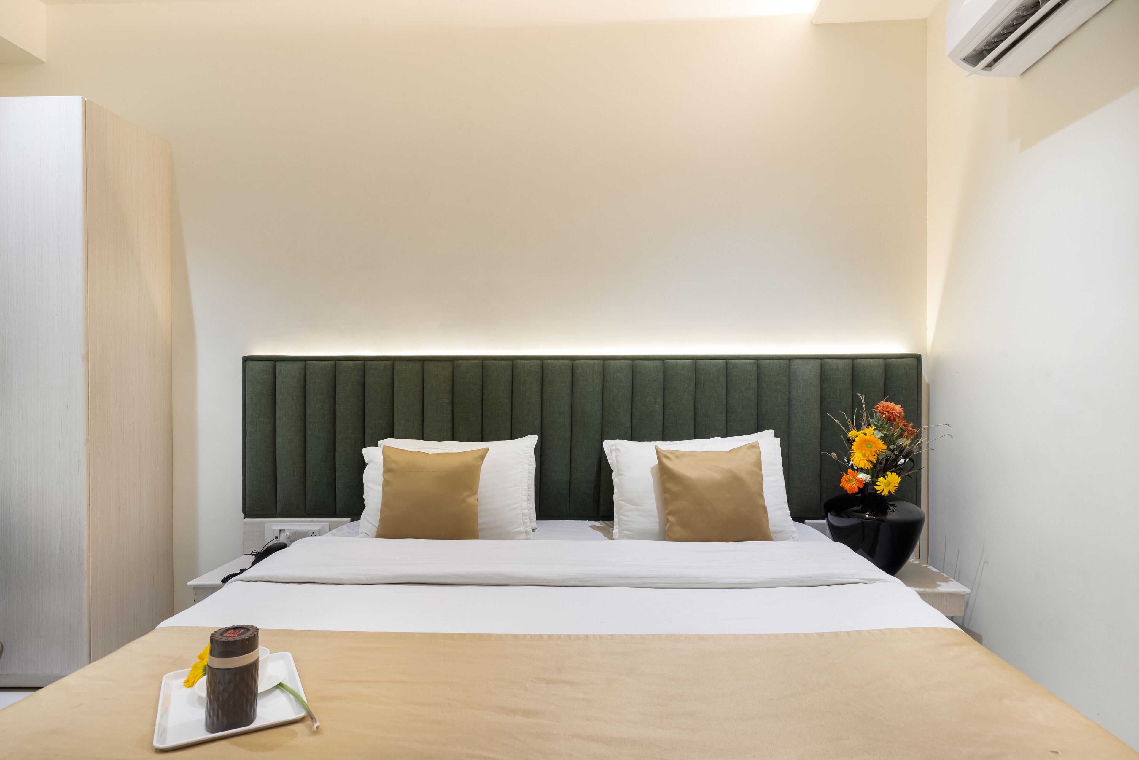 A hotel room with a bed, a bedside table, a nightstand, a wall-mounted air conditioner, and a vase with flowers.