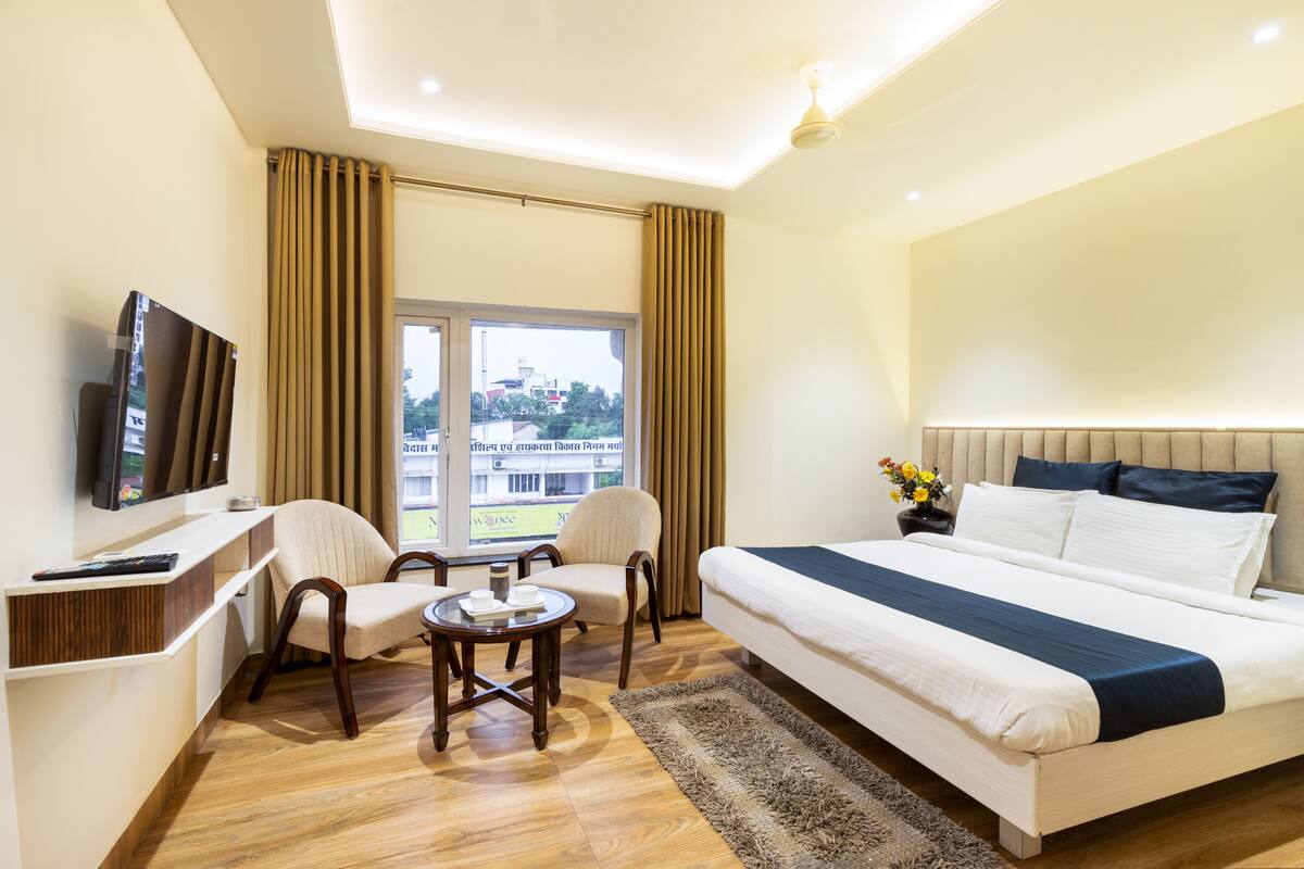Habitación de hotel con cama, televisión, escritorio, dos sillas, una mesita y ventana con cortinas.
