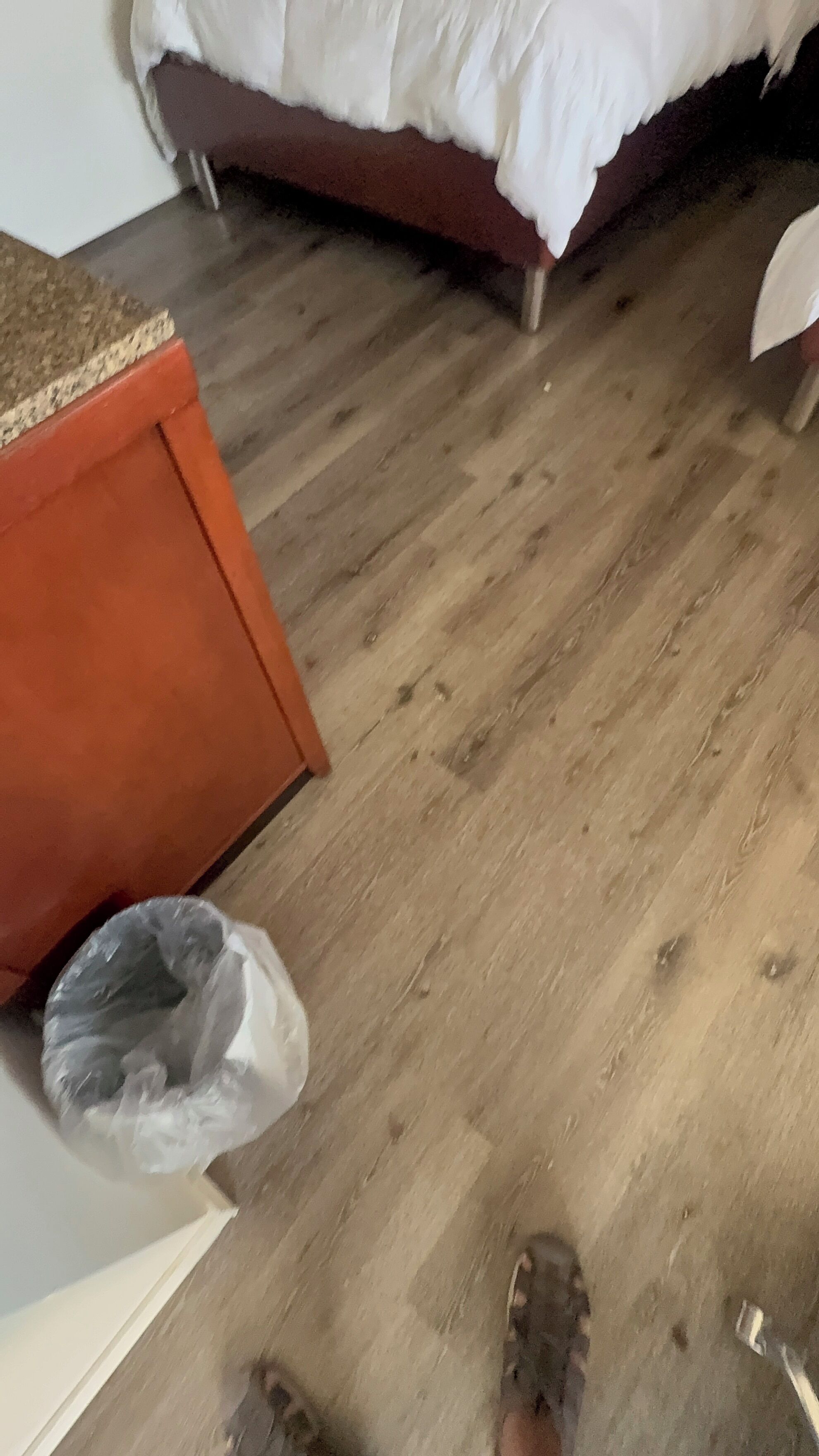 A room with a bed, a wooden floor, a trash bin, and a piece of furniture with a red base.